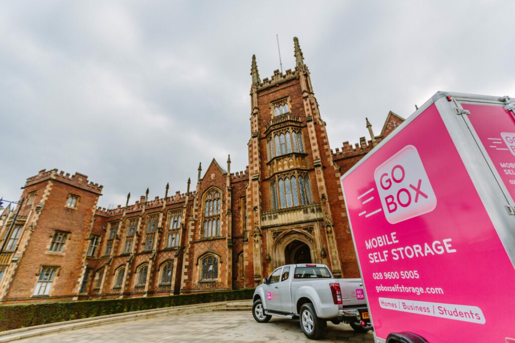 A pick-up truck towing a mobile storage unit in front of a historic gothic-style building under a cloudy sky.