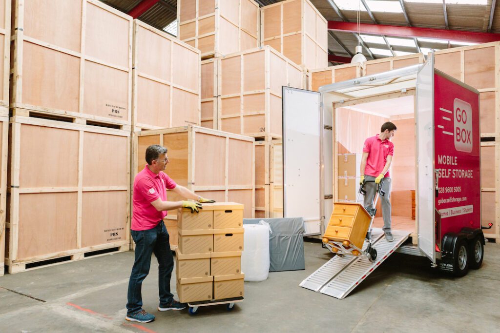 Two people wearing pink shirts are unloading furniture from a mobile storage unit into a self-storage warehouse in Belfast.