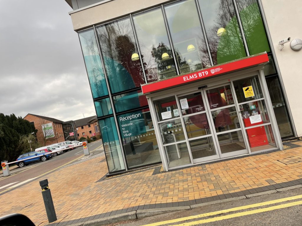 Entrance to elms bt9 with a reception sign, featuring glass doors and a red banner, on an overcast day.