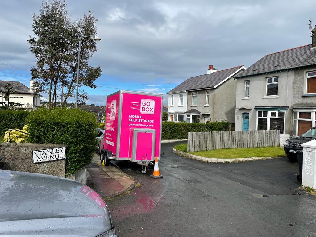 A bright pink mobile Storage Solutions unit parked on a residential street next to semi-detached houses under an overcast sky.