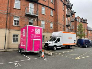 A pink mobile self-storage trailer and a white moving van parked in an urban apartment parking lot.