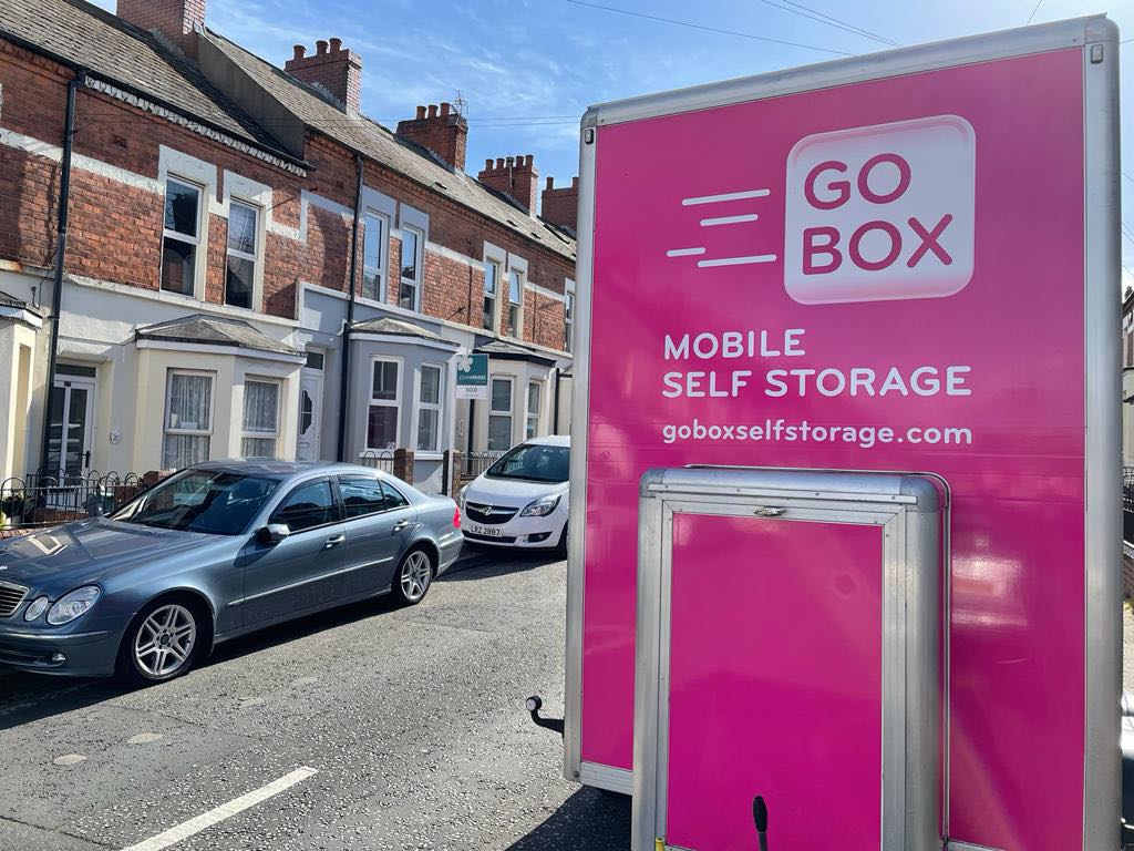 A pink mobile self-storage unit parked on a street with residential houses and cars in the background, offering numerous reasons to choose this convenient option.