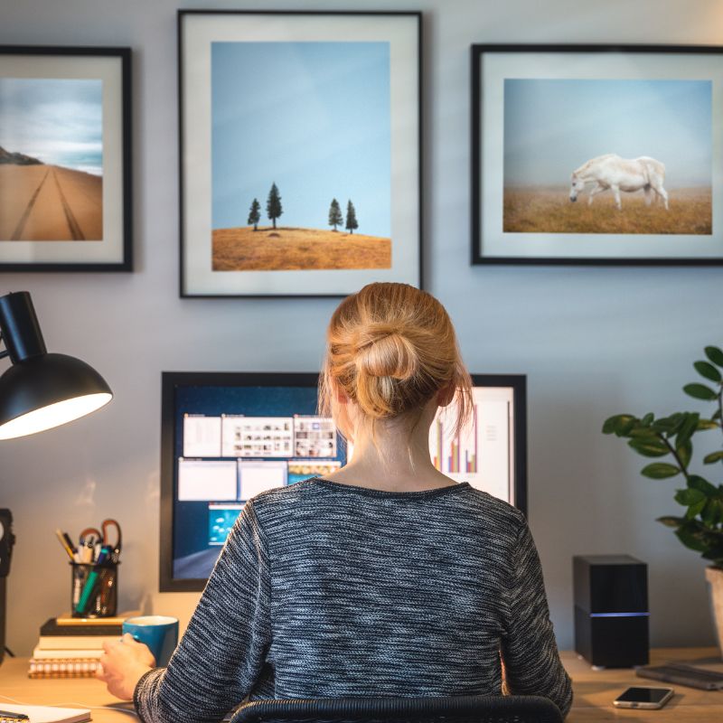 Woman working at a desk with dual monitors, framed pictures on the wall, and a lamp illuminating the workspace.