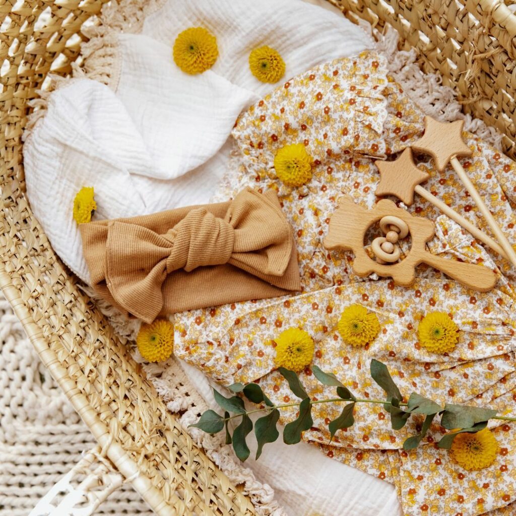 A wicker basket containing baby items: a patterned blanket, a headband, wooden toys, and a sprig of greenery.