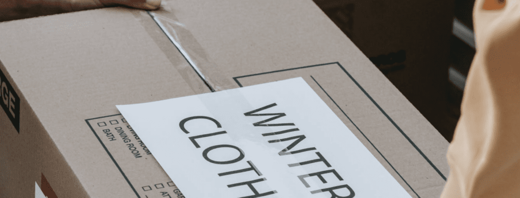 A person carrying a cardboard box labeled 'winter clothes' with a packing tape partially peeled off.