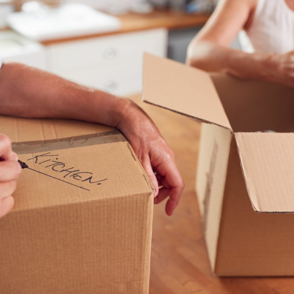 Person labeling a moving box for kitchen items, following the Ultimate Guide for Downsizing.