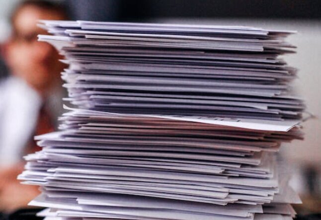 A towering stack of documents on a desk with an out-of-focus person in the background.