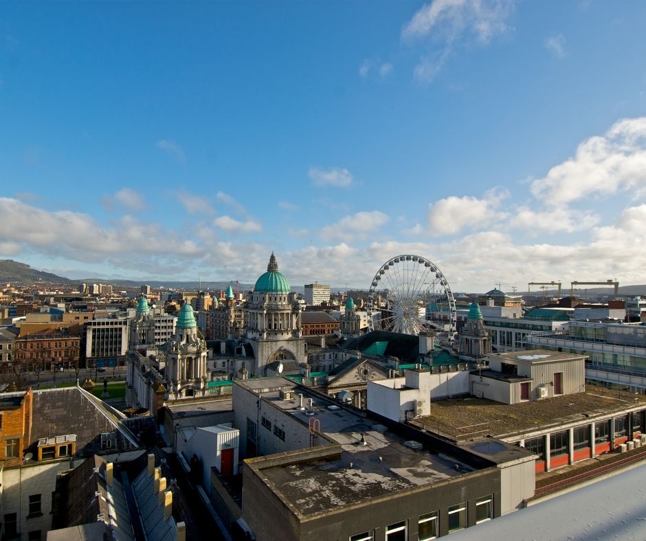 Cityscape with historic buildings and a ferris wheel under a partly cloudy sky.