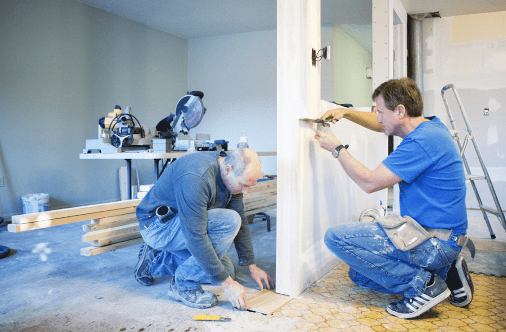 Two men working on a home renovation project, with one installing flooring and the other working on a door frame.