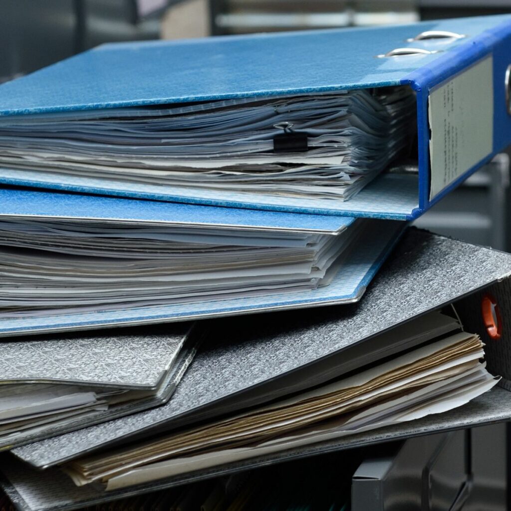 Stacks of binders and documents piled haphazardly on a desk, indicating the potential benefits for offices & businesses of using commercial self storage units.