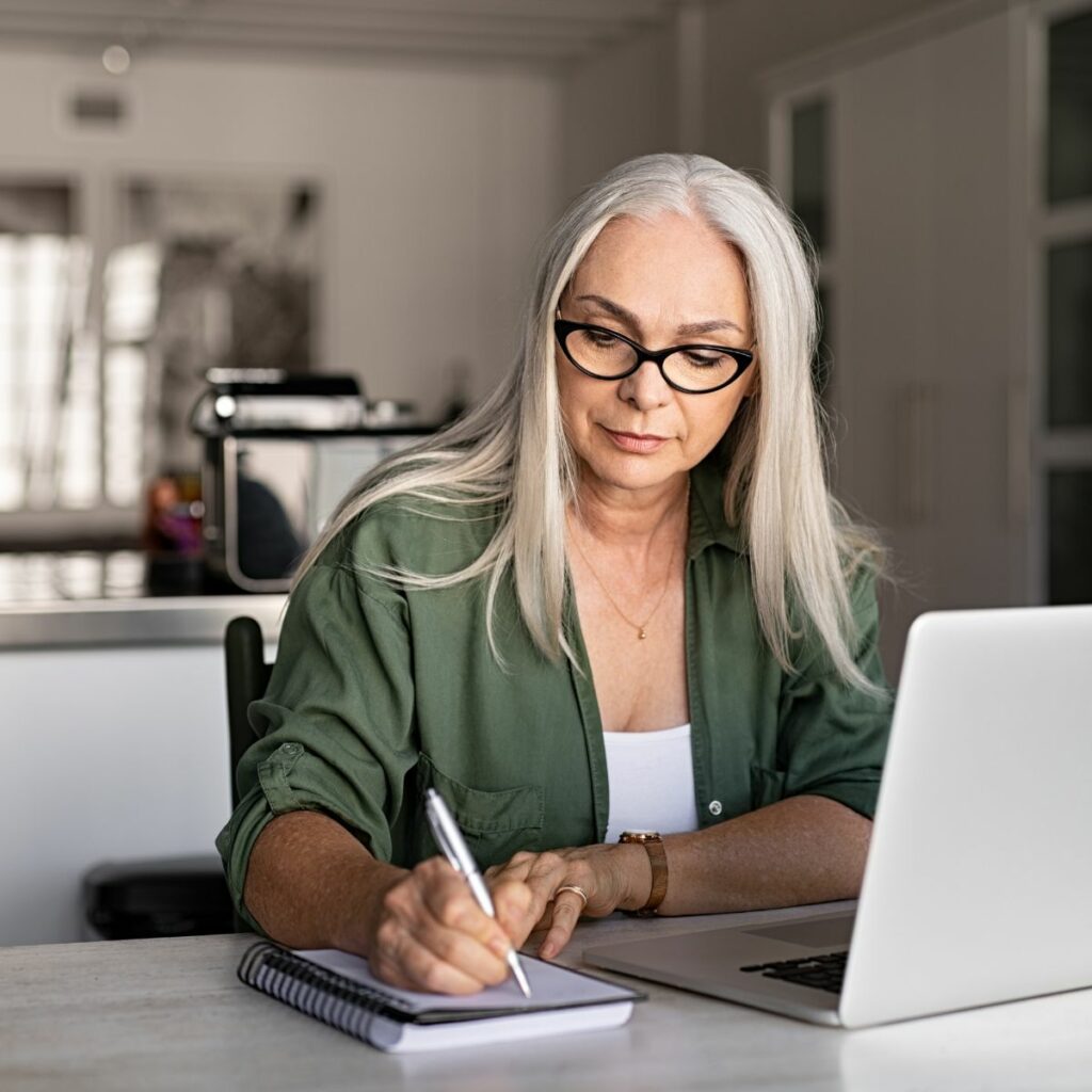 Mature woman with glasses writing notes next to a laptop in a home office setting.