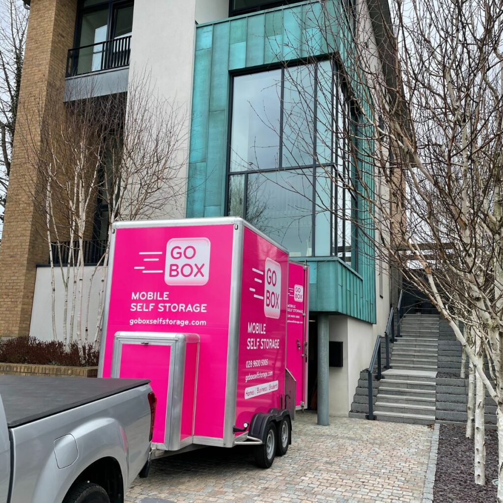 A pink 'go box mobile self storage' trailer parked in front of a modern building with steps and bare trees on either side.