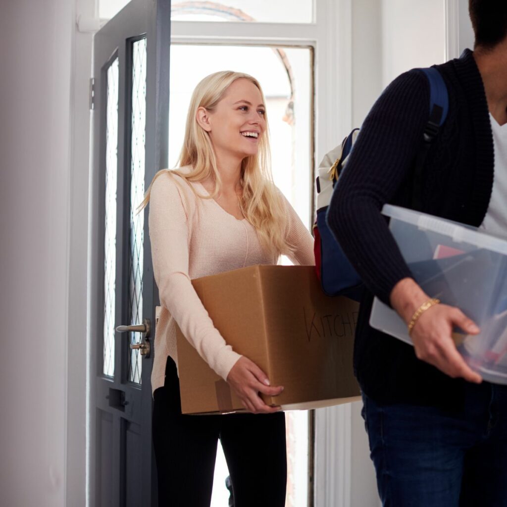 A woman smiling while carrying a cardboard box into a Belfast house, accompanied by an international student holding storage containers.
